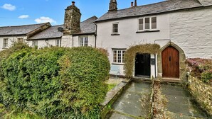 A traditional stone building with a thatched-roof entrance, a narrow stone pathway leading to the door, and a well-maintained hedge in the foreground.