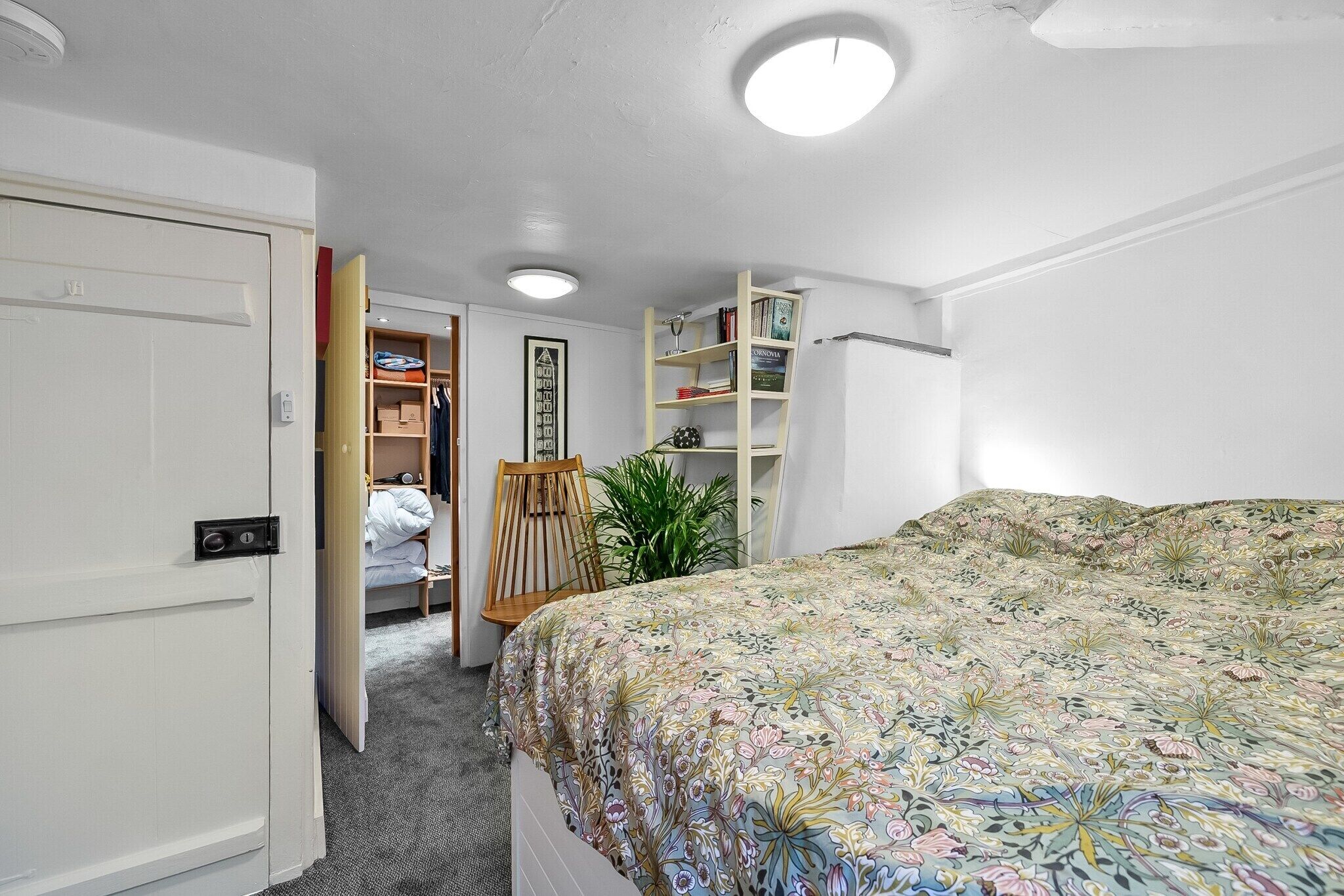 A bedroom with a bed, a wardrobe, a shelf with books, a chair, and a potted plant.