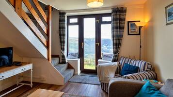 Een gezellige woonkamer met een bank, een salontafel en een televisie op een witte tafel. Er is een trap met houten treden die leidt naar een balkon met uitzicht op het landschap.