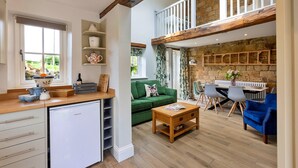 A spacious living area with a green sofa, wooden coffee table, and a dining set. A kitchen area with a white refrigerator and shelves is visible. - Cottage in Grantley near Fountains Abbey (Grantley)