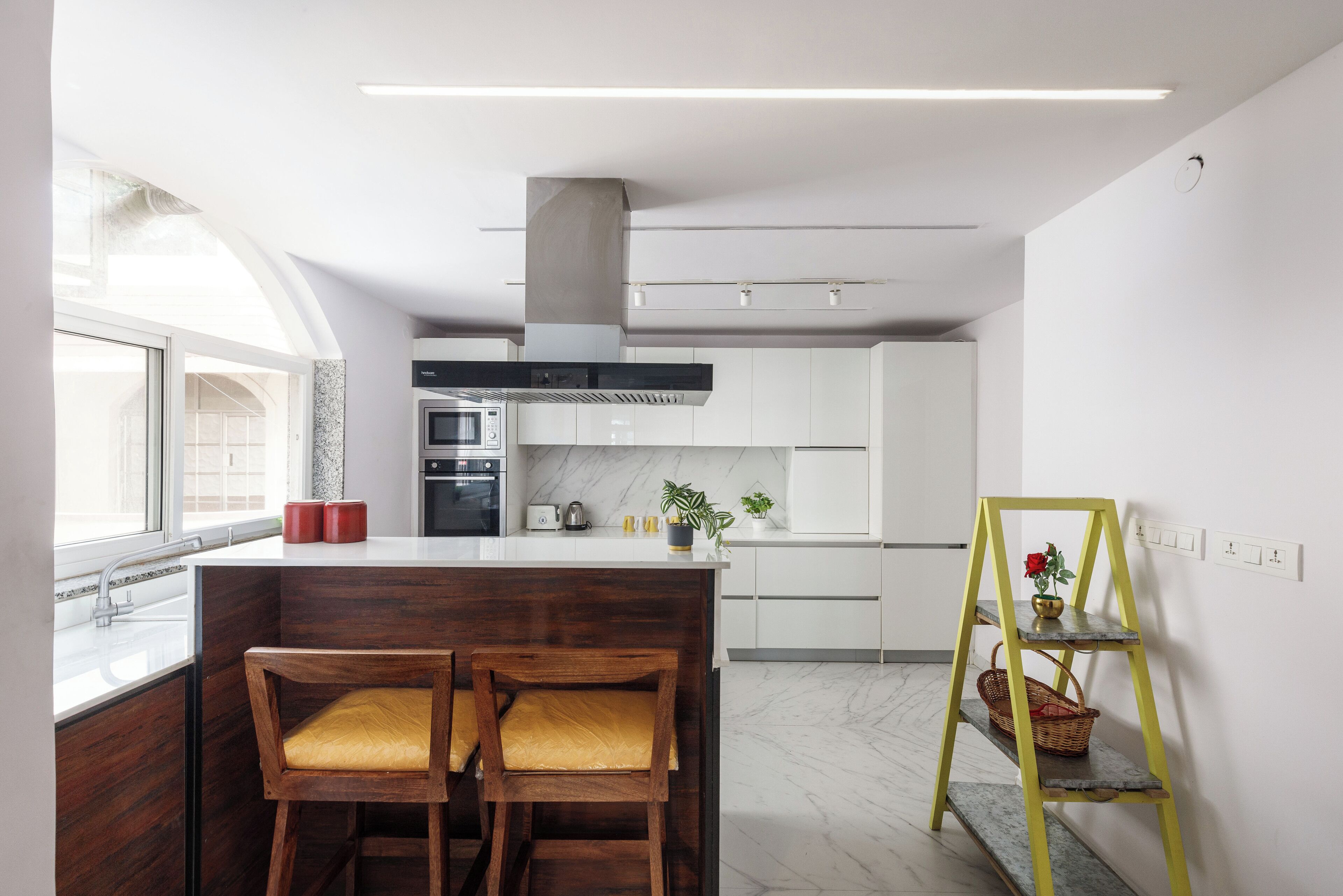 Een moderne keuken met een centraal eiland, houten barstoelen, een gele ladenkast en uitzicht op de stad door grote ramen.
