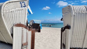 A beachside view with wicker chairs and a striped beach umbrella. - Modern lounge apartment with terrace, beach chair, parking - 50 meters to the beach (Sierksdorf)