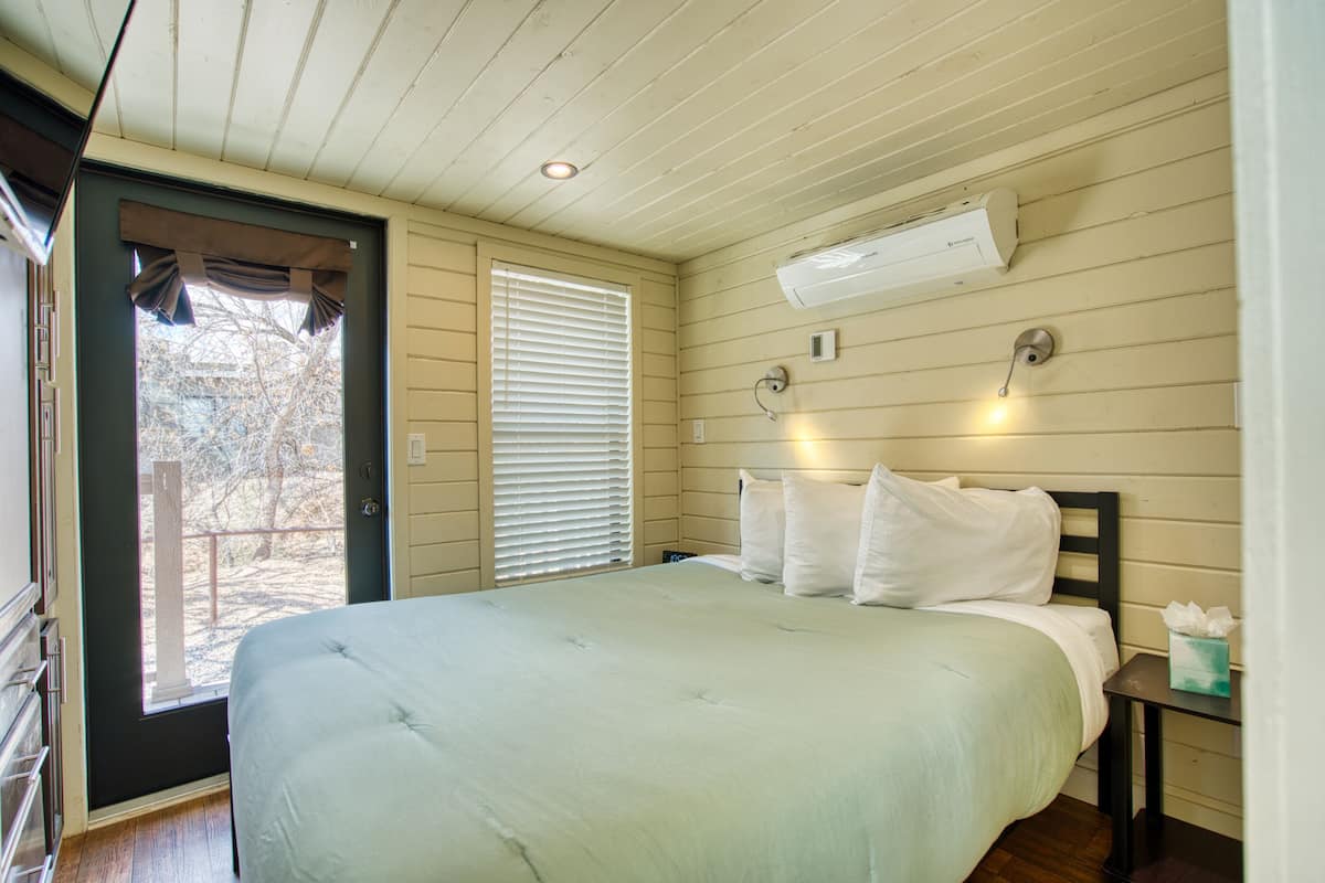 A bedroom with a bed, a door, a window with blinds, an air conditioning unit, and a bedside table with a tissue box.