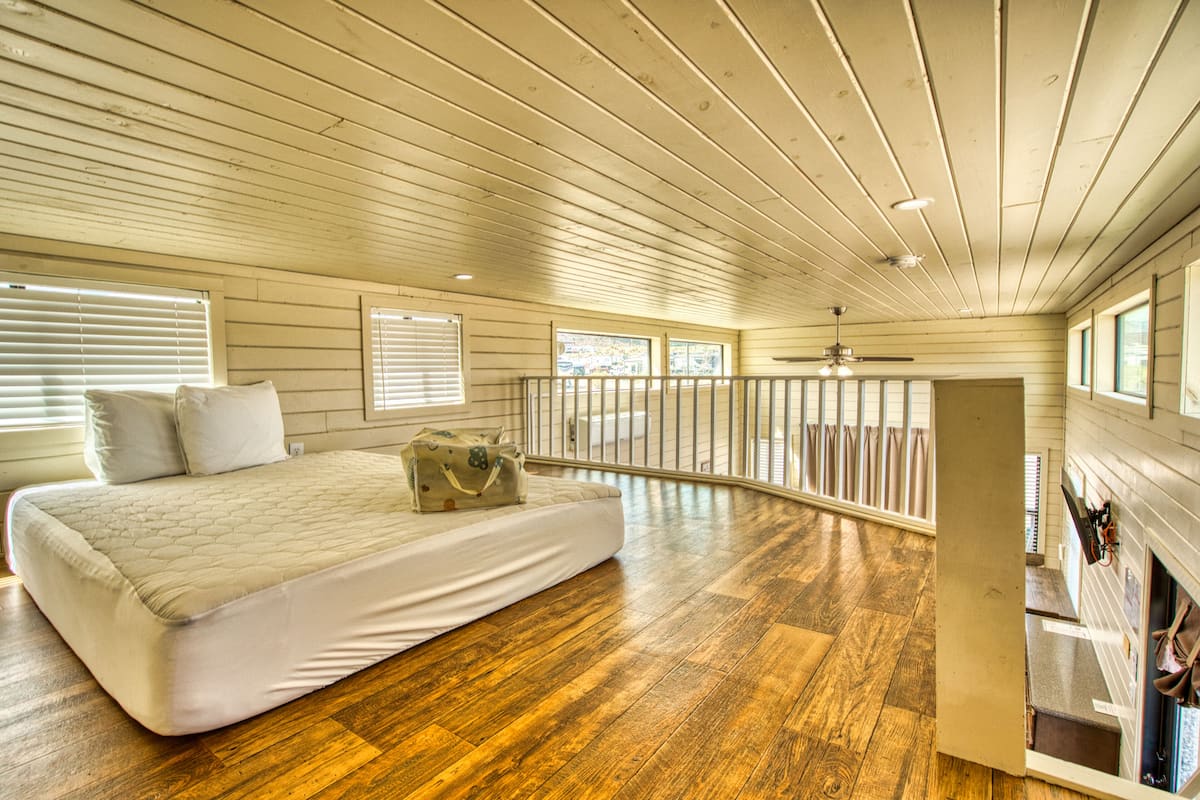 A bedroom with a wooden floor, a bed with pillows, a suitcase, and a balcony with a railing.