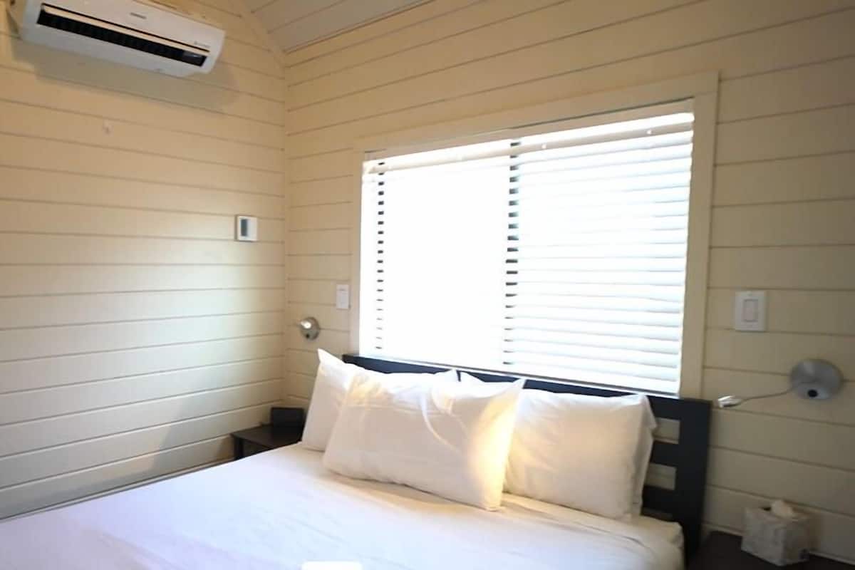 A small, single-bed room with a window, white walls, and a ceiling-mounted air conditioner.