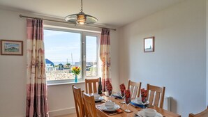 A dining room with a wooden table set for four, a large window with curtains, and a view of a coastal landscape.