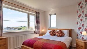 A bedroom with a large window, a bed with a red and pink quilt, a wooden nightstand, and a floral wallpaper on the wall.