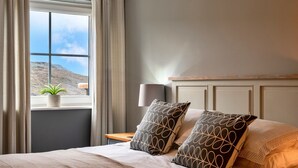 A neatly made bed with patterned pillows, a bedside table with a lamp, and a window with curtains and a potted plant. - Apartment in Watergate Bay with Sea Views (Watergate Bay)