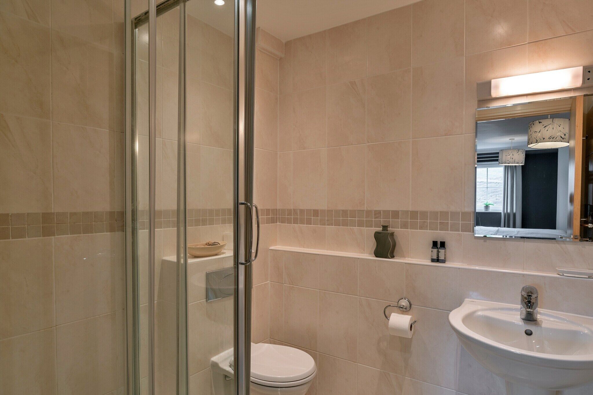 Une salle de bain moderne avec une douche à parois de verre, des toilettes blanches, un lavabo avec un miroir au-dessus et une étagère contenant des articles de toilette.