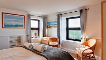 A hotel room with a bed, two wicker chairs, a dresser, a window with curtains, and a painting on the wall.