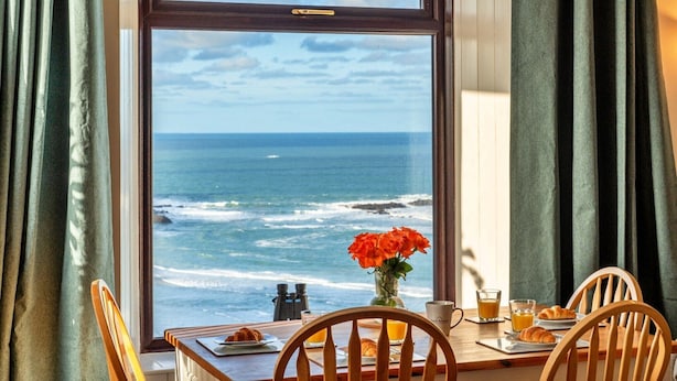 A dining table with chairs, a vase with flowers, a plate with food, a glass of juice, and a view of the ocean through a window.