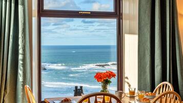 A dining table with chairs, a vase with flowers, a plate with food, a glass of juice, and a view of the ocean through a window.