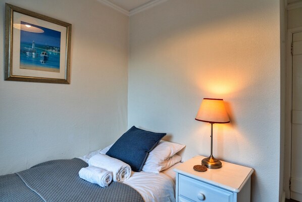 A neatly made bed with a blue pillow, a white bedside table with a lamp, and a framed picture of a coastal scene on the wall.