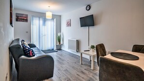 Een moderne woonkamer met een bank, een eettafel en stoelen. Er is een televisie aan de muur bevestigd, een plant op de tafel en een raam met jaloezieën.