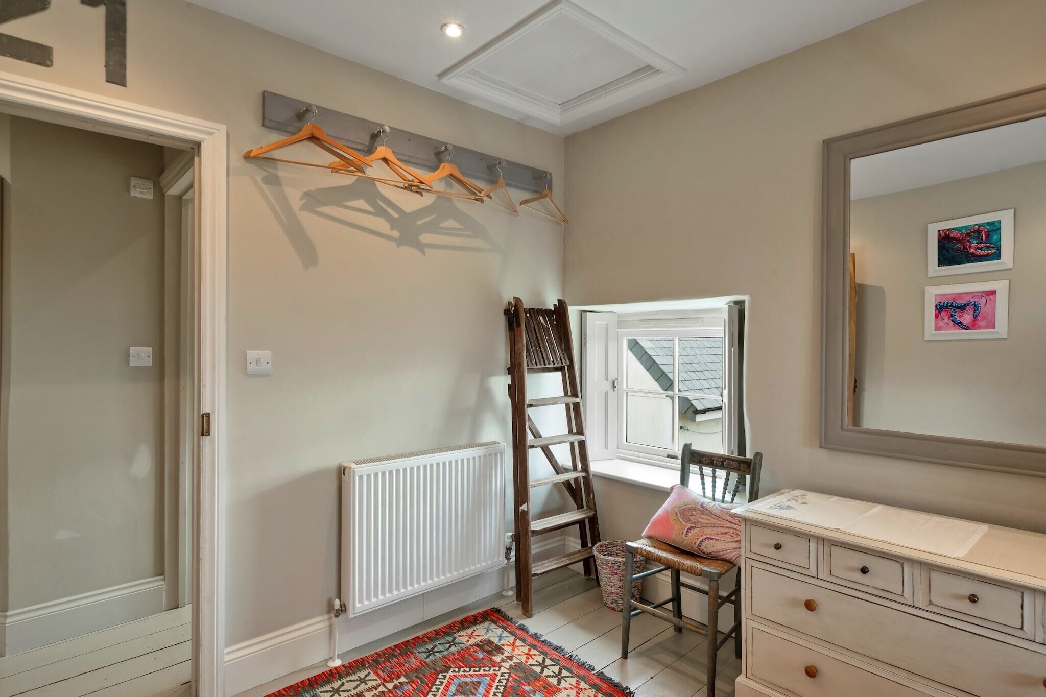 A room with a white dresser, a chair with a cushion, a radiator, a wooden ladder, and a window with a view.