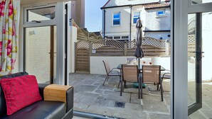 A patio area with a table and chairs, a sofa with a red cushion, and a view of a house through a glass door.
