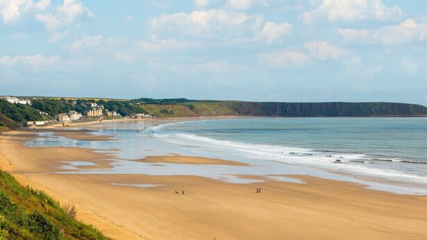 A coastal beach with a sandy shore, a few people, and a building complex in the background.
