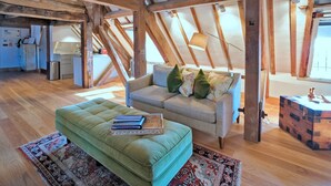 A loft-style living space with exposed wooden beams, a sofa with decorative pillows, a green ottoman with books, and a rustic wooden chest. - Apartment in Brackley Town Hall Charm (Brackley)