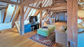 A loft-style room with exposed wooden beams, a television on a wooden stand, a green ottoman, a patterned rug, and a sofa. - Apartment in Brackley Town Hall Charm (Brackley)