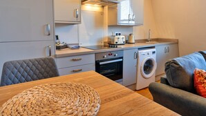 A compact kitchen with a toaster, oven, and washing machine. There is a wooden table with a woven placemat and a grey sofa with a cushion. - Apartment in Matlock near Peak District Trails (Matlock)