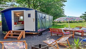 Une petite maison mobile bleue et blanche, dotée d’un lit et d’une fenêtre, entourée de meubles de jardin et d’un parasol coloré.
