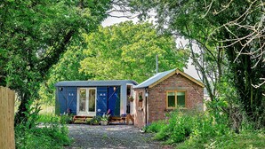 Une petite cabane rustique aux murs en briques, avec une structure bleue attenante, le tout entouré d’une végétation luxuriante.