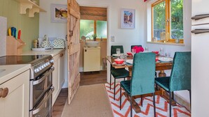 Une cuisine compacte équipée d’une cuisinière, d’un évier et de meubles en bois. Un coin repas avec une table pour quatre personnes, des chaises vertes et une fenêtre donnant sur un paysage verdoyant.