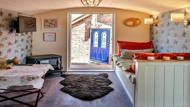 Une pièce chaleureuse avec un plafond en bois, une porte bleue, un lit avec des oreillers rouges, une petite table recouverte d’une nappe blanche et une cheminée.