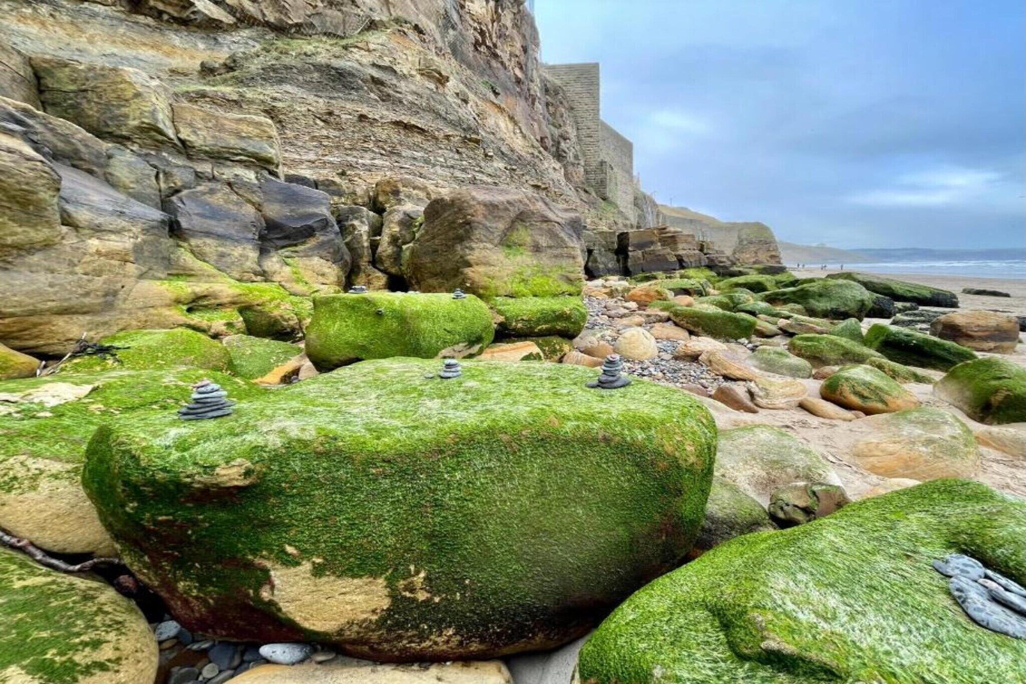 Una spiaggia rocciosa con grandi massi ricoperti di muschio e una scogliera sullo sfondo.
