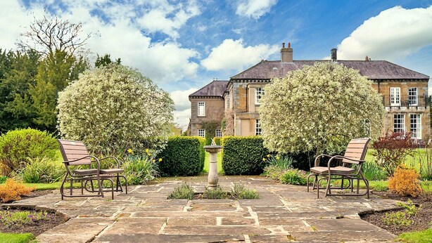 A stone pathway leading to a large house with two symmetrical trees and benches on either side.