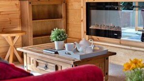 Interior acogedor de una cabaña de madera, con una mesa de madera dispuesta con tetera blanca, tazas y una pequeña planta en maceta. Se ve una chimenea al fondo.