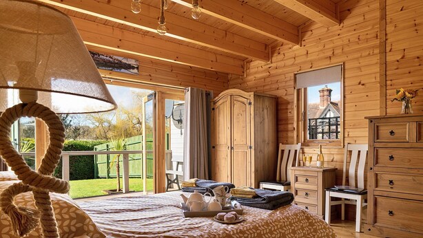 Una habitación de cabaña de madera con una cama, un aparador, una silla y una mesa con una botella y vasos.