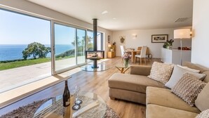A spacious living room with a large window overlooking the sea, a fireplace, a sofa, and a dining area.