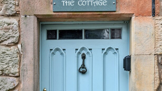 Une petite maison avec une porte bleu clair, un heurtoir en laiton et une enseigne au-dessus de la porte sur laquelle on peut lire : « THE COTTAGE ».