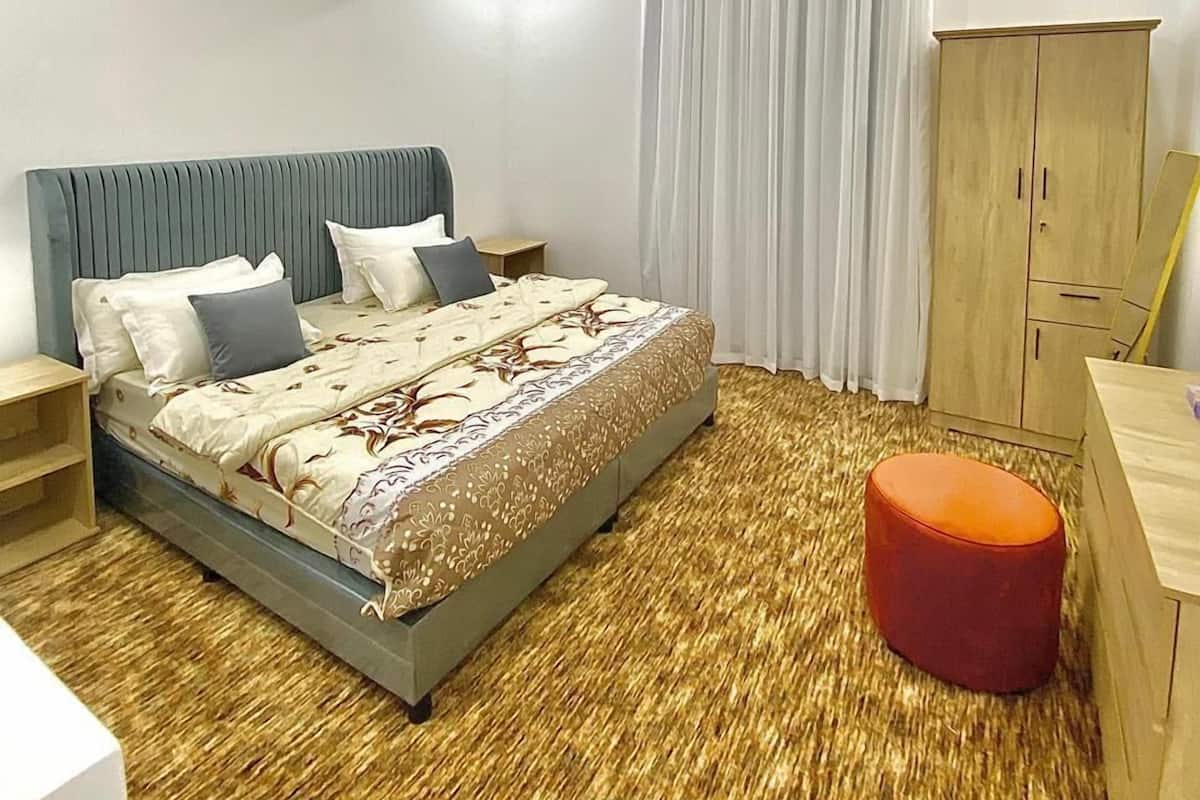 A modern bedroom with a large bed, a wooden wardrobe, a small table, and an air conditioning unit.