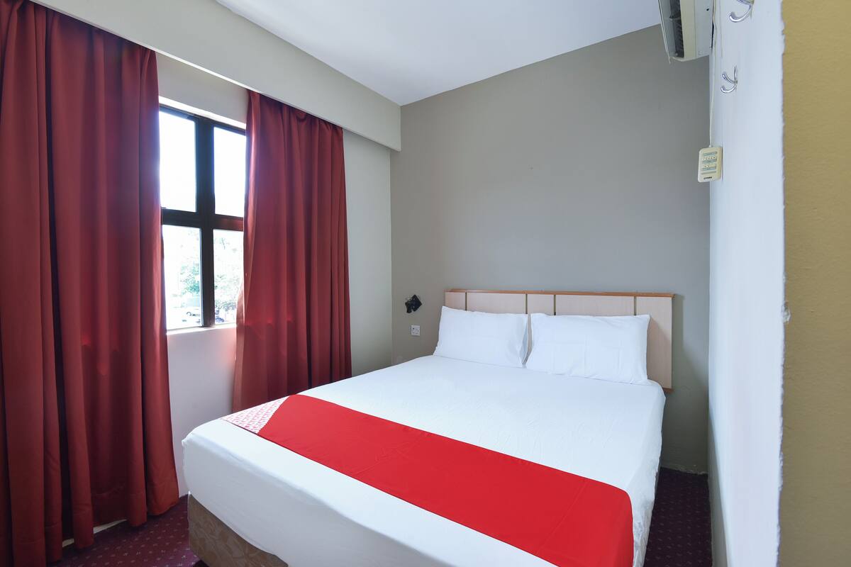 A hotel room with a bed, red curtains, a window, and a wall-mounted air conditioning unit.