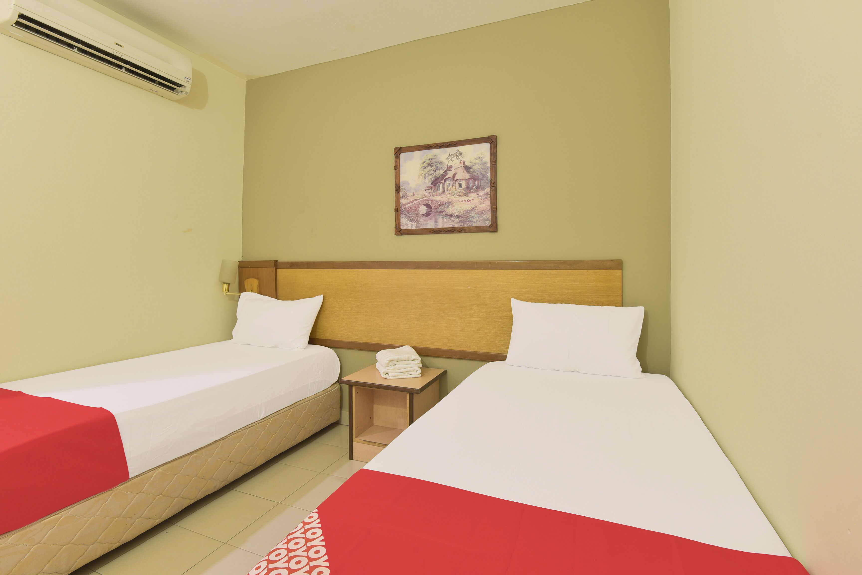 Habitación de hotel con dos camas individuales, cabecero de madera, una mesita de noche con lámpara y un equipo de aire acondicionado en la pared.