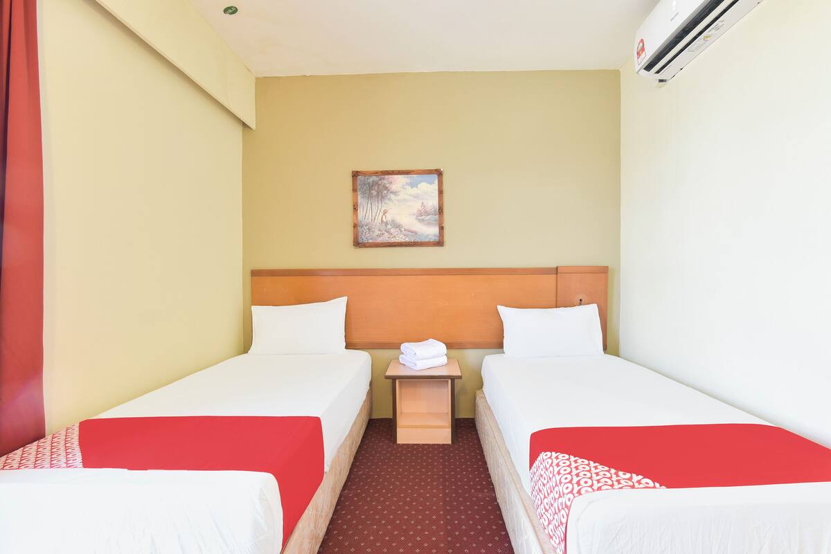 A hotel room with two beds, a small wooden nightstand, a painting on the wall, and an air conditioning unit.