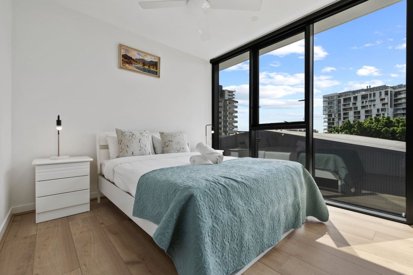 A modern bedroom with a large bed, a bedside table, and a view of a cityscape through floor-to-ceiling windows.