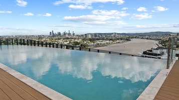 A rooftop infinity pool with a city view, wooden deck, and a clear sky.