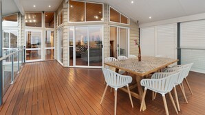 Una amplia terraza con suelo de madera, una mesa de comedor con sillas y un ventanal que da a un espacio interior.