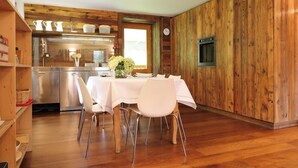 A dining area with a wooden wall, a kitchen in the background, and a clock on the wall. - Apartment in a chalet nestled in the forest, with sauna and private forest (Canal San Bovo)