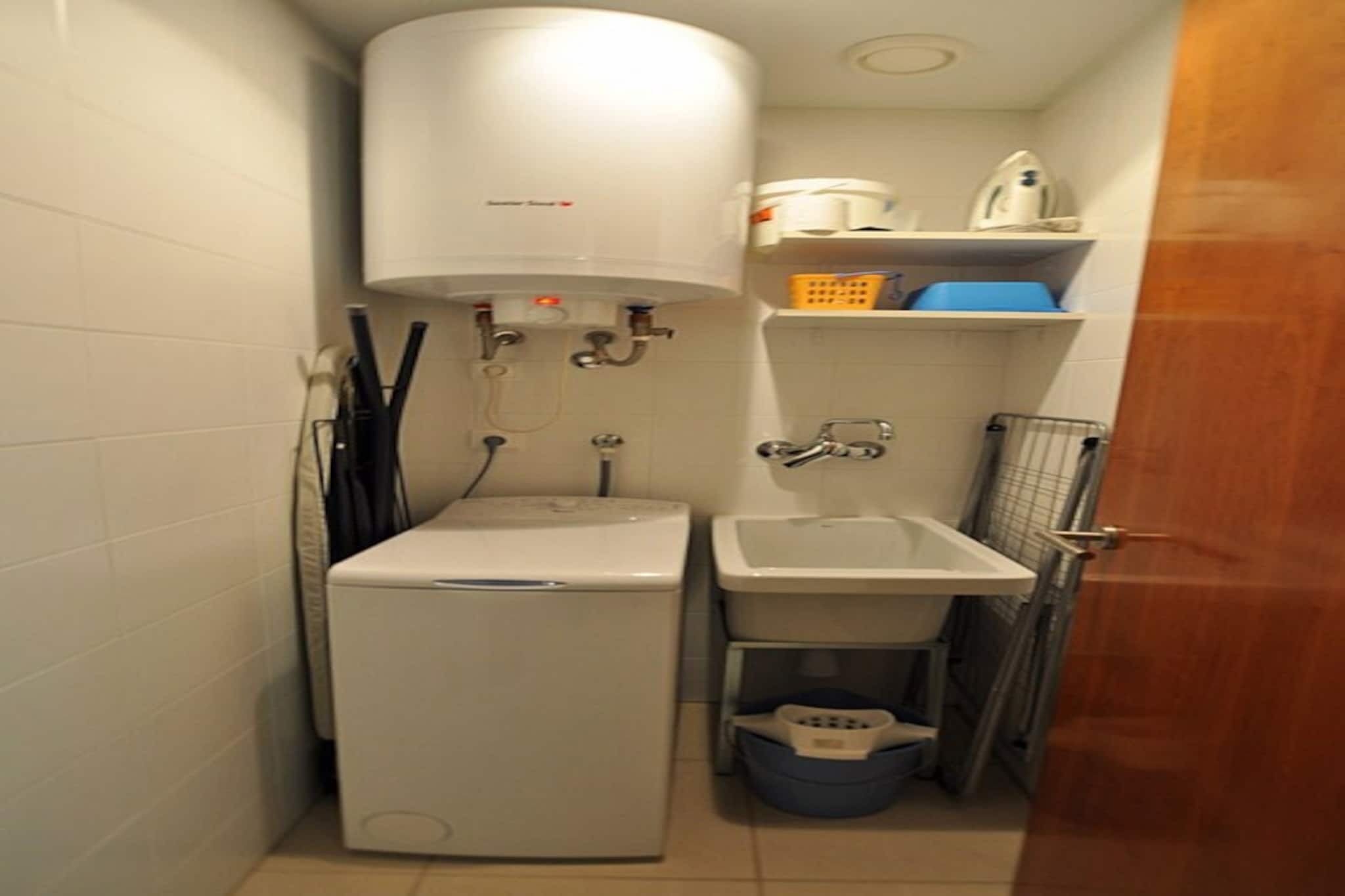 A compact bathroom with a white toilet, sink, and a water heater. There is a blue laundry basket and a metal rack.