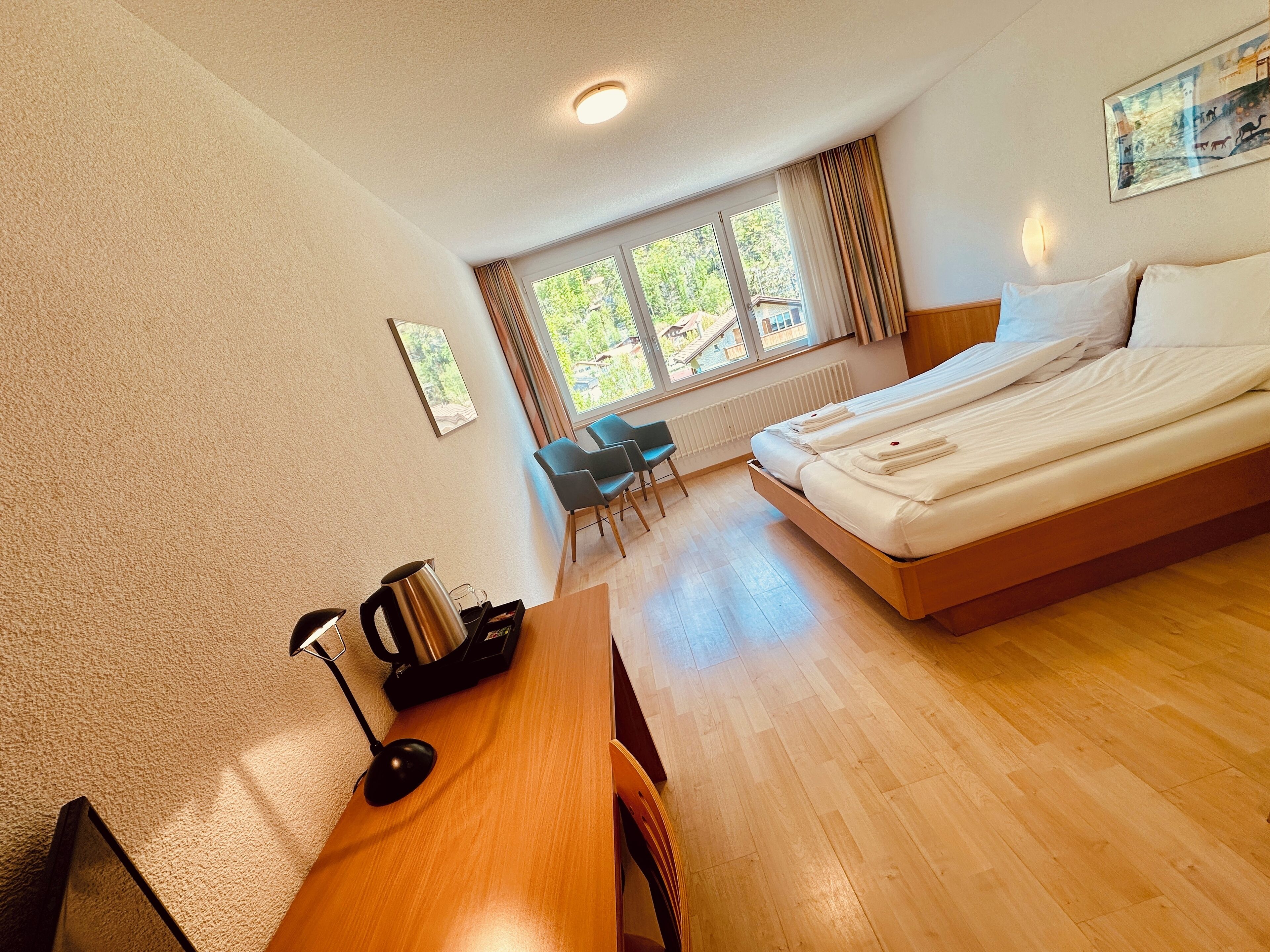 Quarto de hotel com uma cama grande, uma escrivaninha com chaleira, uma cadeira e uma janela com cortinas.