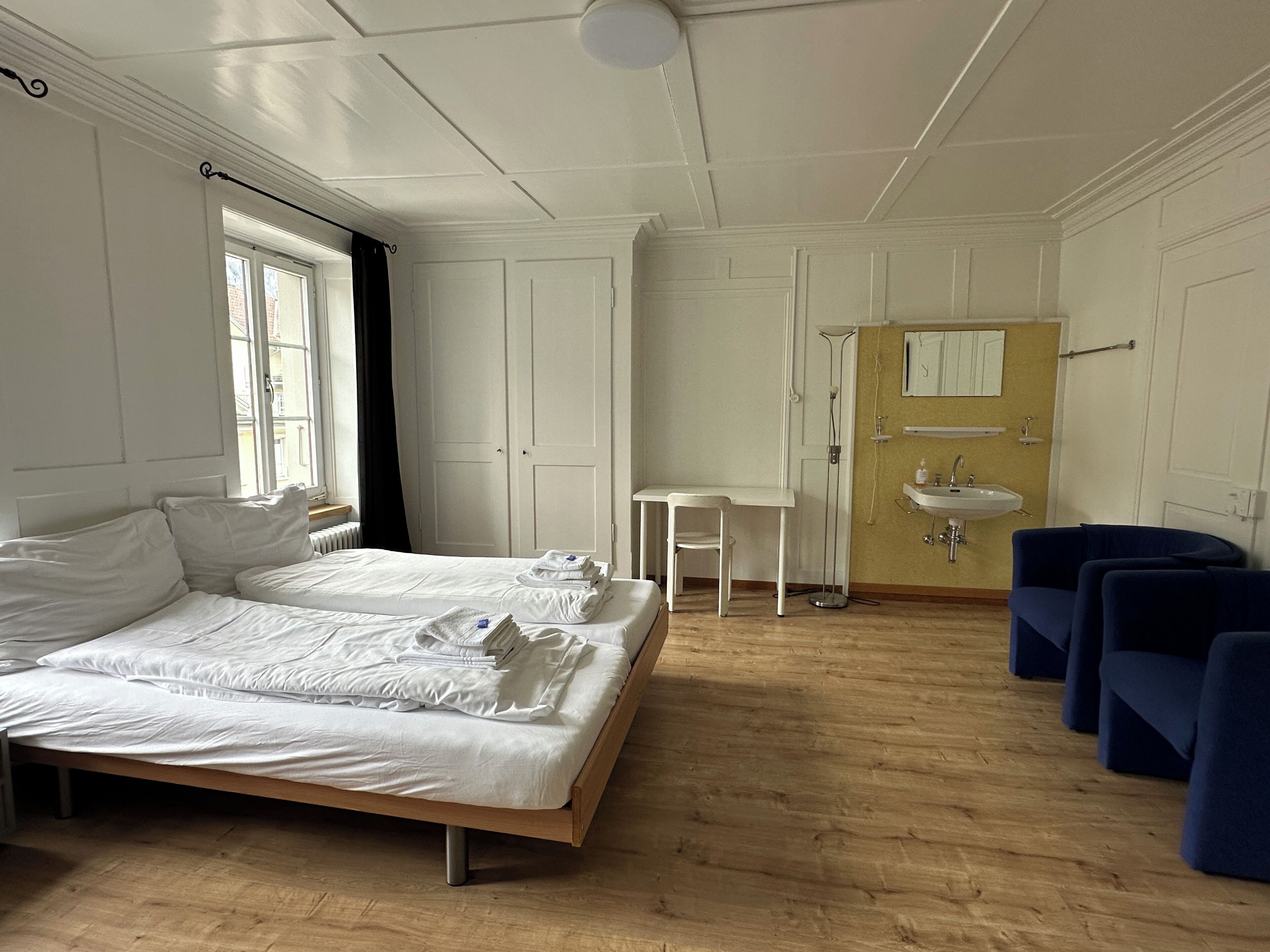 Una habitación de hotel con cama, una mesita, una silla, un lavamanos y un espejo.