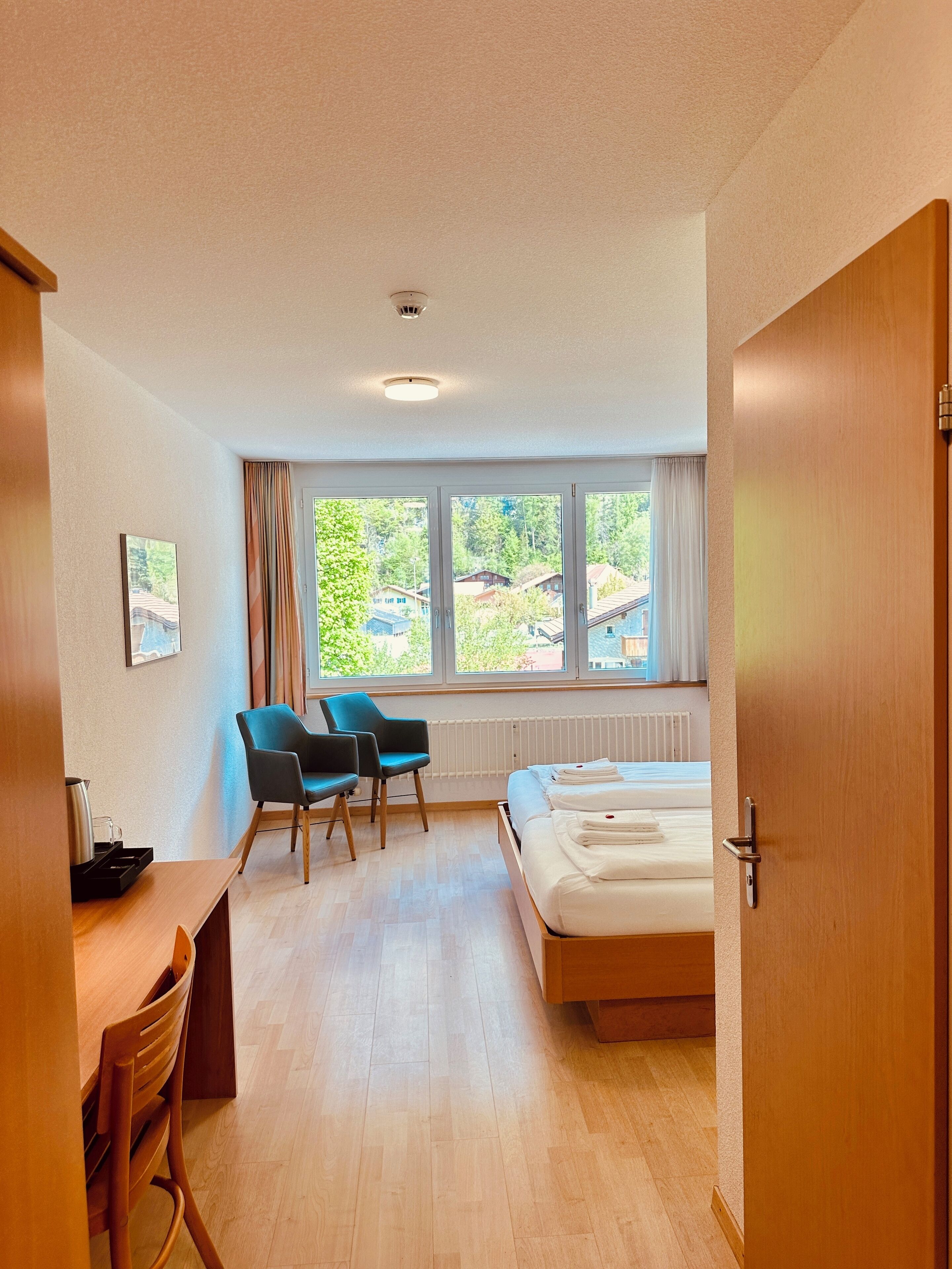 A modern hotel room with a bed, a desk, two chairs, and a large window offering a view of the outdoors.