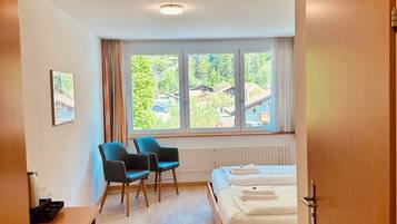 A modern hotel room with a bed, a desk, two chairs, and a large window offering a view of the outdoors.