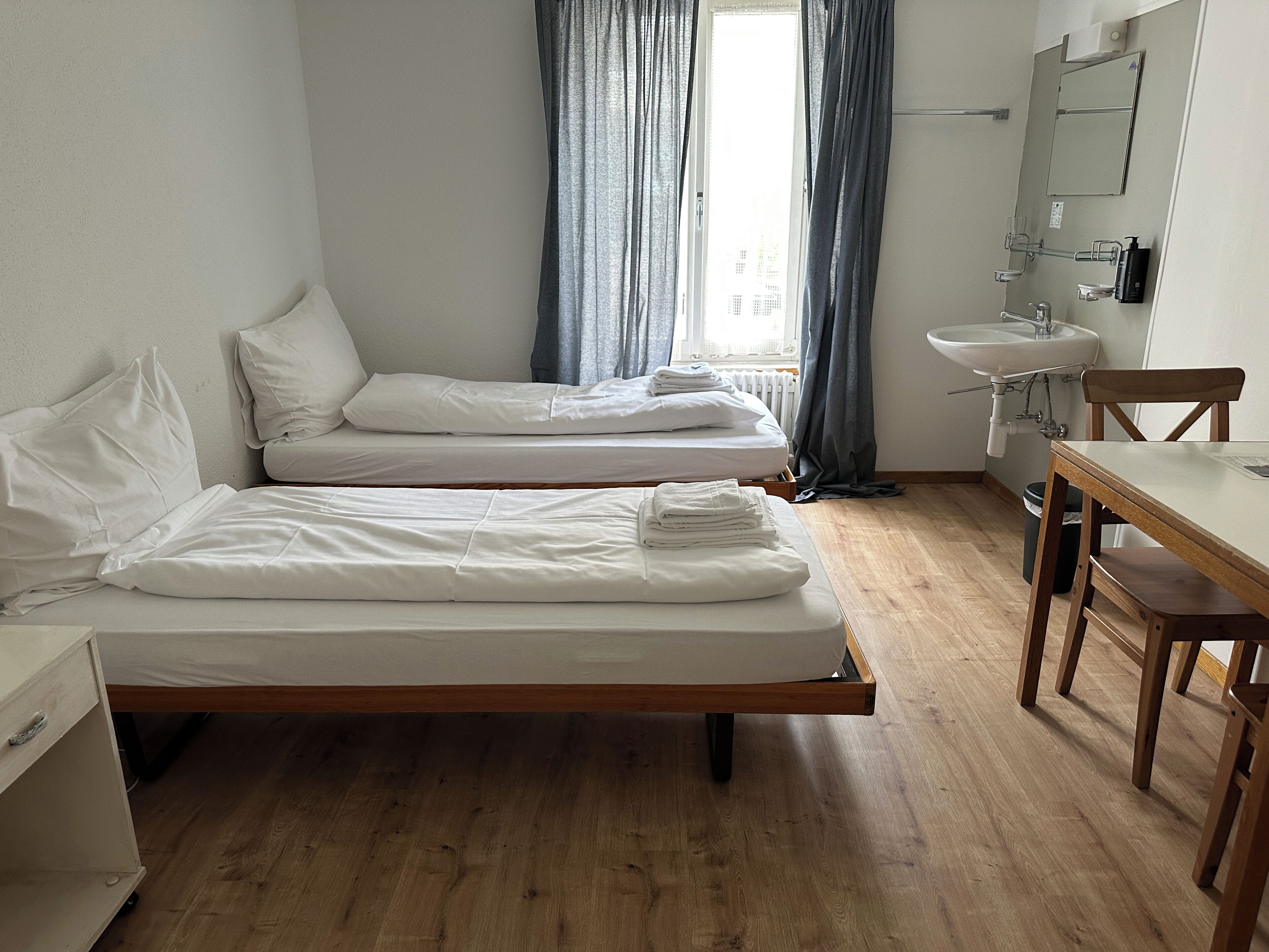 Una habitación con dos camas, una mesa de madera, una silla, un lavamanos y una ventana con cortinas.