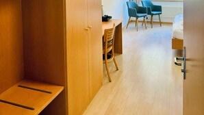 Ein Hotelzimmer mit einem Holzschrank, einem Schreibtisch mit zwei Stühlen, einem Bett und einem Fenster mit Blick ins Grüne.
