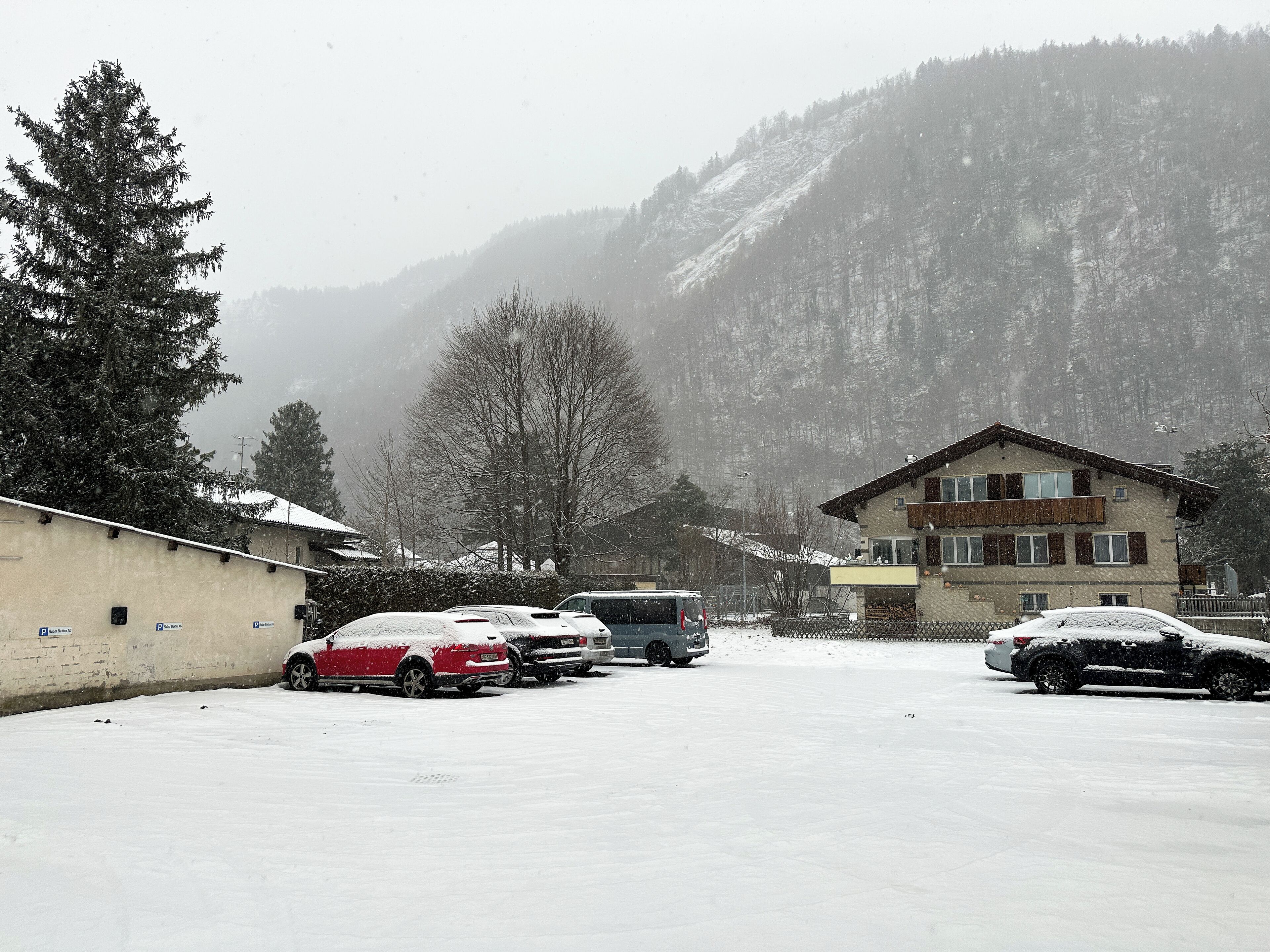A snow-covered parking lot with several cars and a bus, a building with a sloped roof, and a mountain in the background.